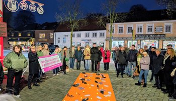 Aufnahme der Mahnwache zum Orange-Day. Zu sehen sind Menschen um einen orangenen Teppich, der durch Texte und Gegenstände auf Gewalt gegen Frauen hinweist.