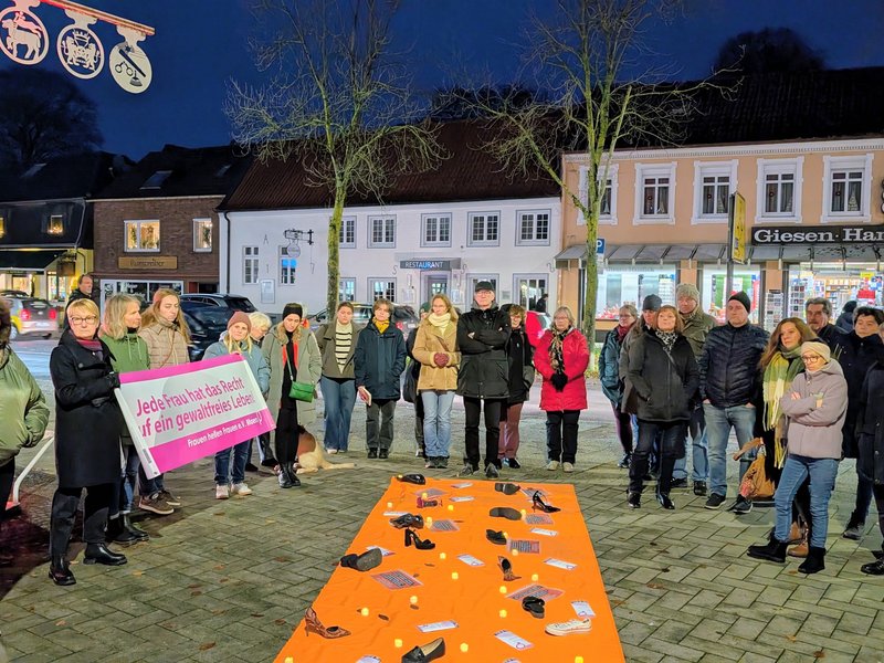 Aufnahme der Mahnwache zum Orange-Day. Zu sehen sind Menschen um einen orangenen Teppich, der durch Texte und Gegenstände auf Gewalt gegen Frauen hinweist.