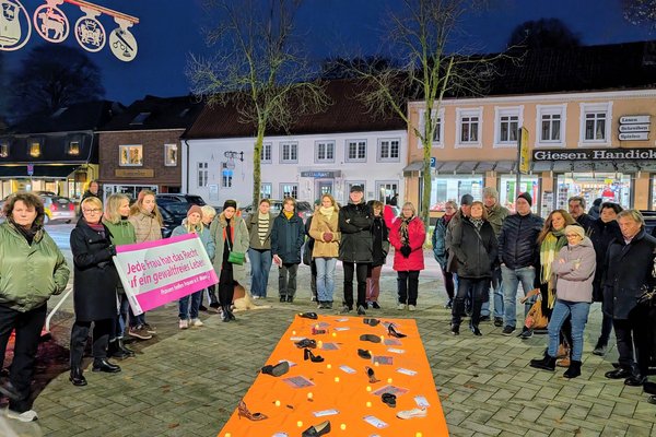Aufnahme der Mahnwache zum Orange-Day. Zu sehen sind Menschen um einen orangenen Teppich, der durch Texte und Gegenstände auf Gewalt gegen Frauen hinweist.